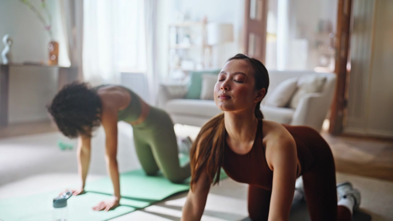 Women doing cat cow yoga stretch home. Fitness friends training together indoor.