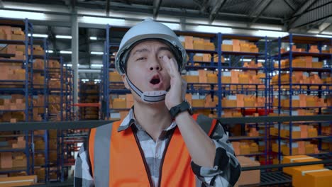 close up of asian male engineer with safety helmet yelling with hand over mouth while standing in the warehouse with shelves full of delivery goods
