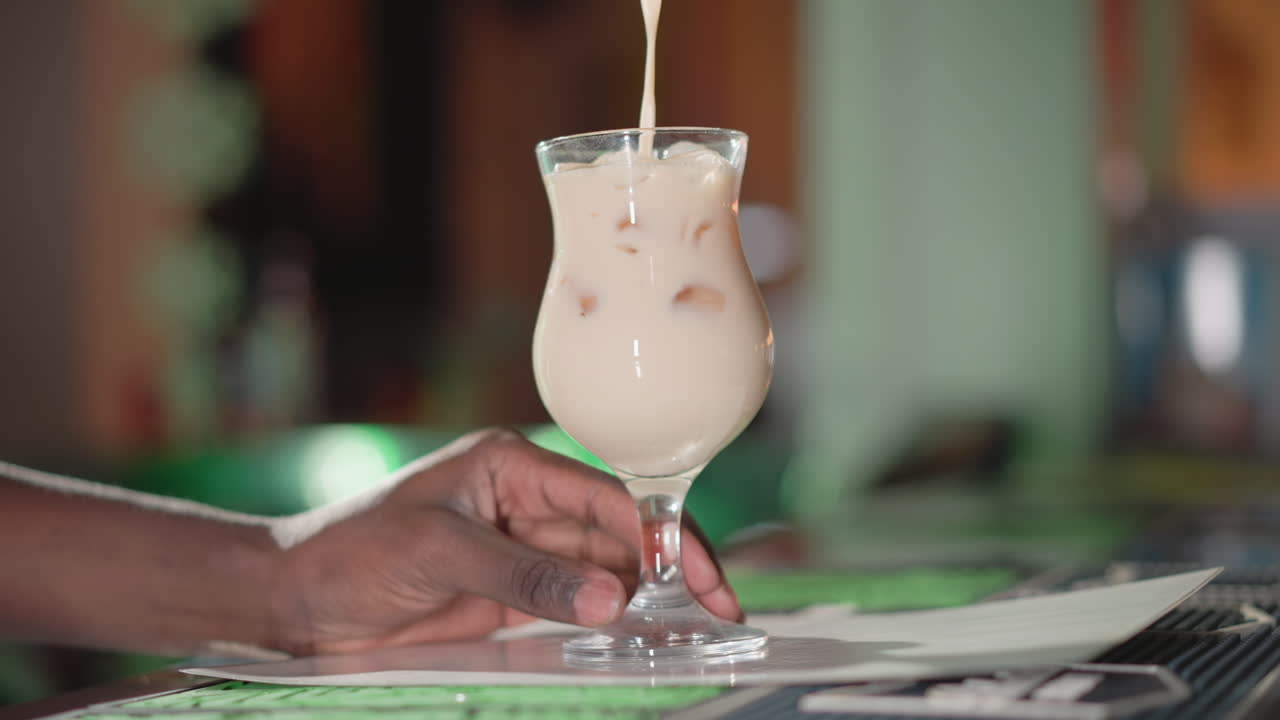 Bartender pouring creamy liquid into glass filled with ice, focusing on the hand and smooth liquid flow in neon-lit bar environment