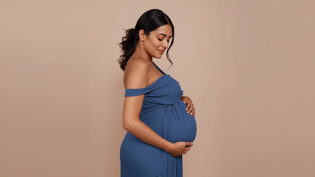 Feeling baby move, pregnant woman in blue dress cradling belly, hair tied, in beige studio