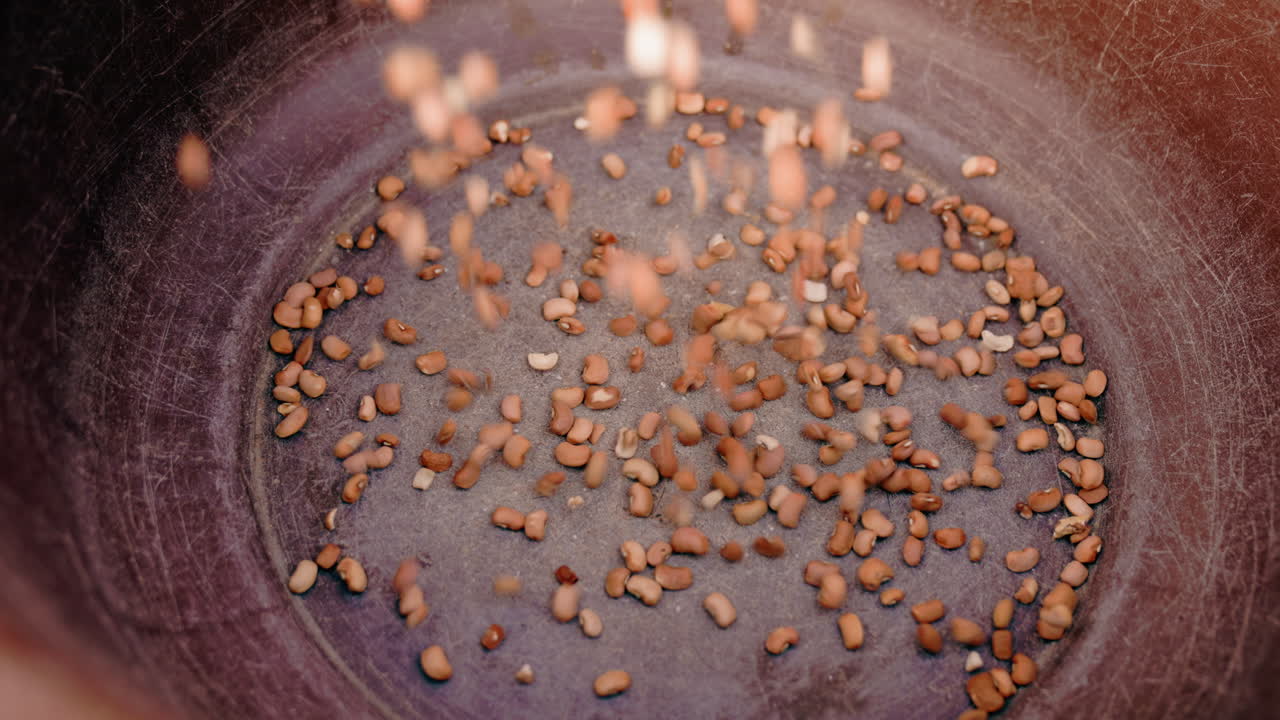 Frigideira preta com grãos sendo despejados, em um close-up de pequenos grânulos marrons caindo em uma panela temperada, acumulando-se gradualmente no centro, sobre uma superfície de metal texturizada, em tons quentes e terrosos, evocando um ritual tátil e calmo.