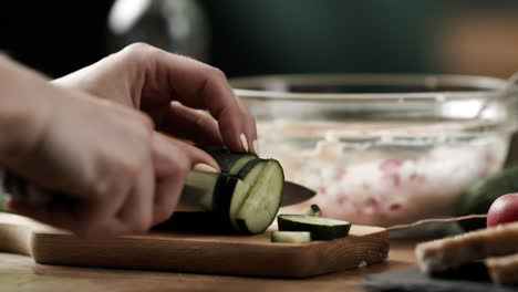 cerca de un cocinero aficionado cortando pepino fresco en trozos pequeños para preparar verduras para untar en la mesa de la cocina de madera.