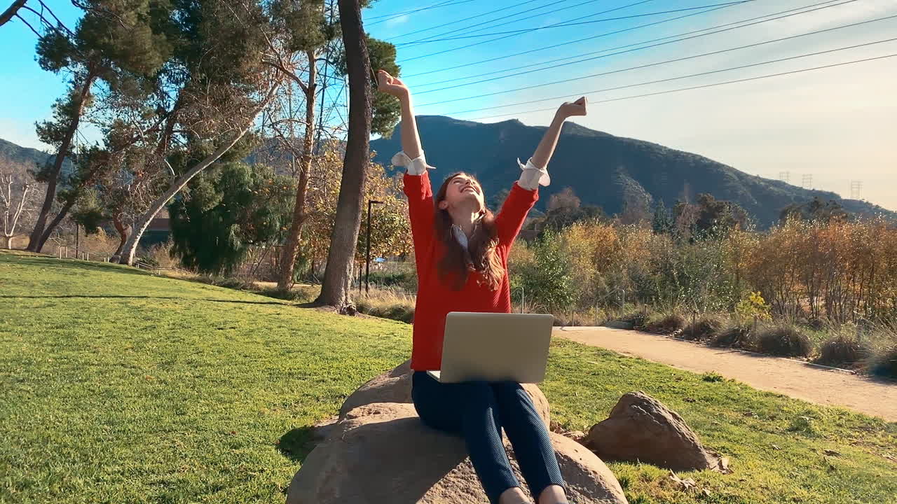 A happy, attractive, young, caucasian woman on her laptop at the park, throws her arms in the air to celebrate her achievement