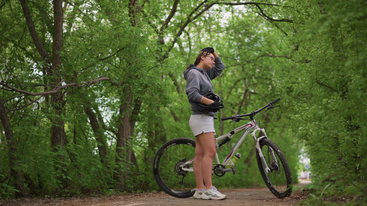 Asian Cyclist Taking Reflective Pause In Green Tunnel, Holding Helmet To Chest And Listening To Birds, Soft Shaded Canopy And Winding Dirt Track, Peaceful Nature Lover Moment Before Ride