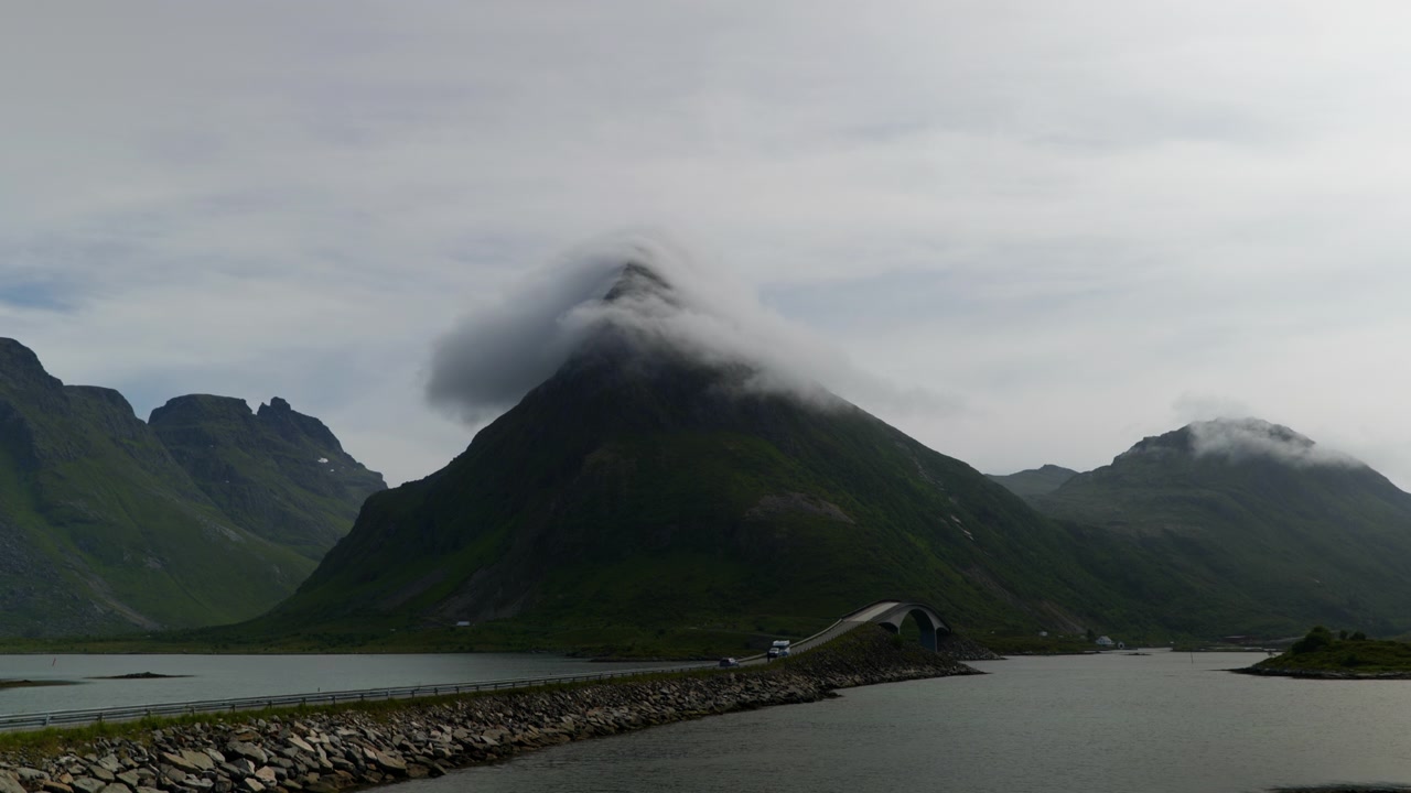 Nuvole sottili avvolgono una ripida cima montuosa verde mentre una strada costiera curva e un ponte attraversano acque calme nelle Lofoten, Norvegia