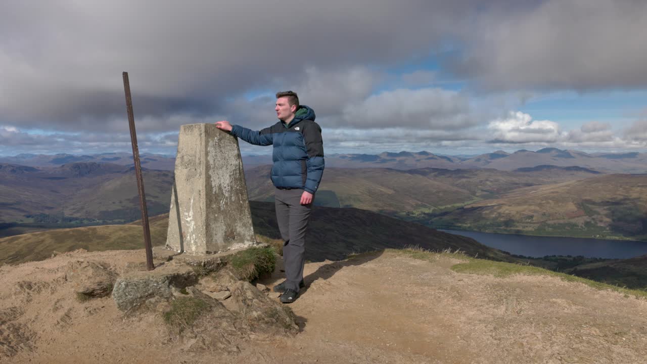 Static shot of a hiker bagging Ben Vorlich touching the trig point and walking