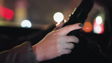 first-person view of a woman's hand with black painted nails tapping on a steering wheel at night. the background features colorful, blurry city lights