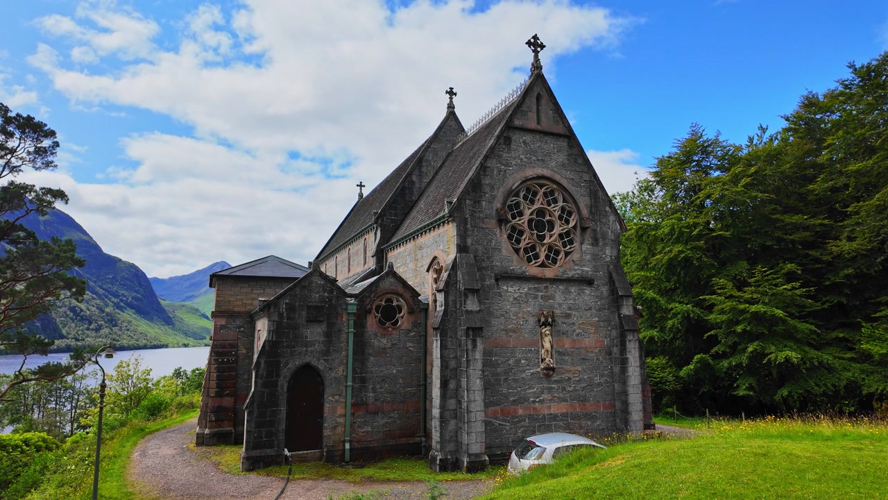 Den idylliske Saint Mary og Saint Finnan kirke i Glenfinnan, Skottland, med Loch Shiel og vakre fjell i bakgrunnen på en solrik sommerdag.