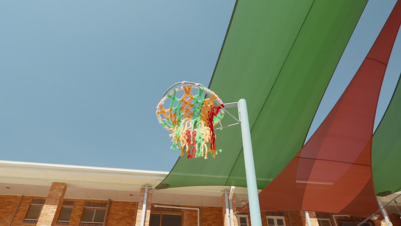 A basket ball being thrown and missing the colourful basketball net with the blue sky and green and red shade sails in the background