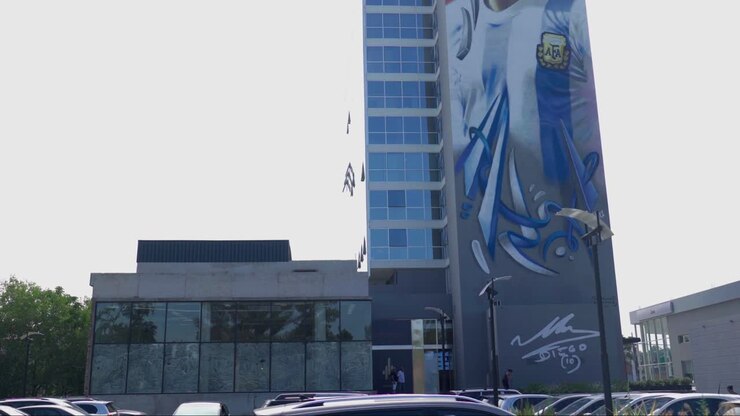 Uma cena em movimento capturando a partida do estacionamento, com o impressionante mural de Maradona dominando a paisagem de fundo