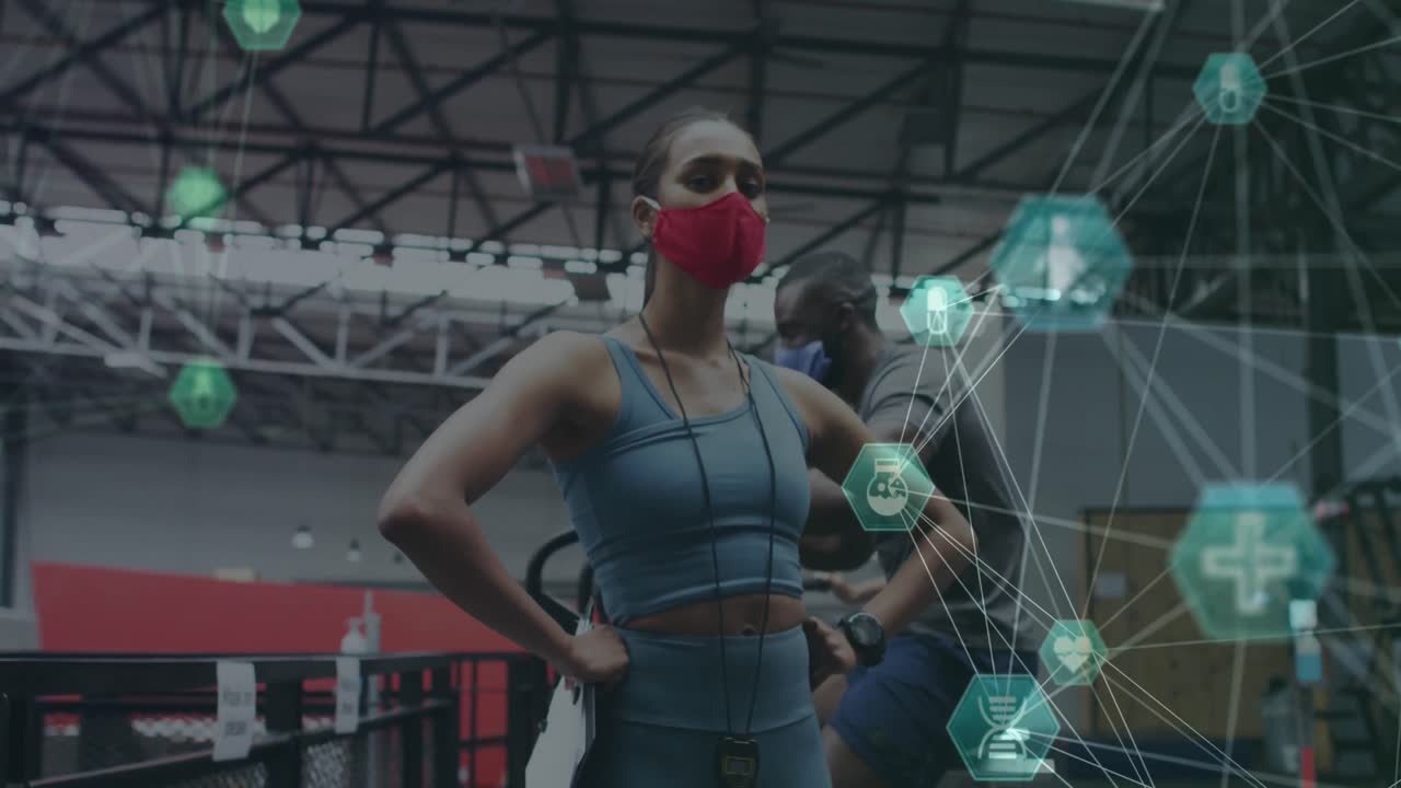 체육관 에 서 있는 얼굴 마스크 를 착용 한 여성 피트니스 트레이너 를 상대로 회전 하는 의료 아이콘 의 세계