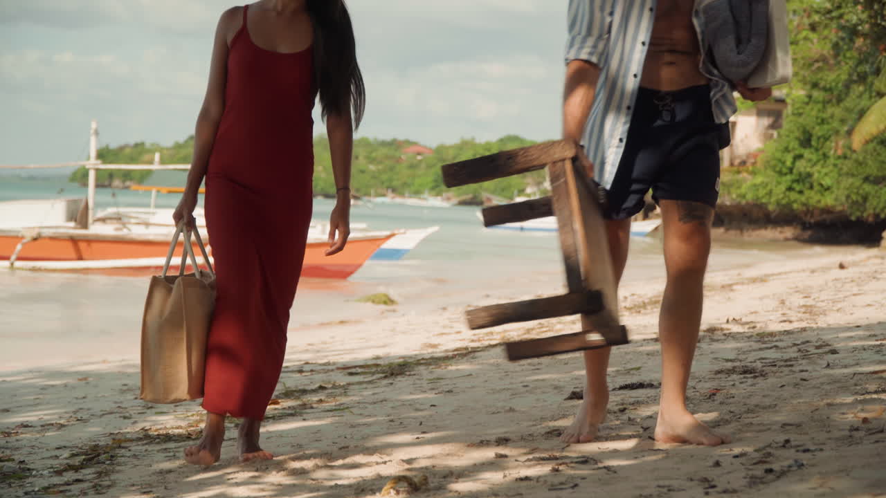 Coppia che cammina sulla spiaggia portando una sedia, posizionando uno sgabello rustico in legno accanto alla barca, donna in vestito rosso che tiene una borsa di paglia, uomo con cappello che porta una sedia pieghevole, impronte a piedi nudi nella sabbia, palme e delicata