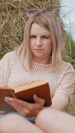 mujer sentada con las piernas unidas, profundamente absorta en la lectura mientras se apoya en la bala de heno, expresión pensativa mientras da la vuelta a las páginas, con un suave borrón en el fondo de los árboles y el campo abierto