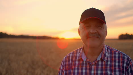 portrait of a senior adult farmer in a field of grain looking at the camera and smiling at sunset. the tractor driver takes off his cap and looks at the camera in slow motion