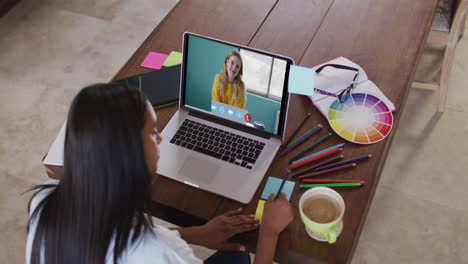 mujer caucásica usando una computadora portátil en una videollamada con una colega, tomando notas
