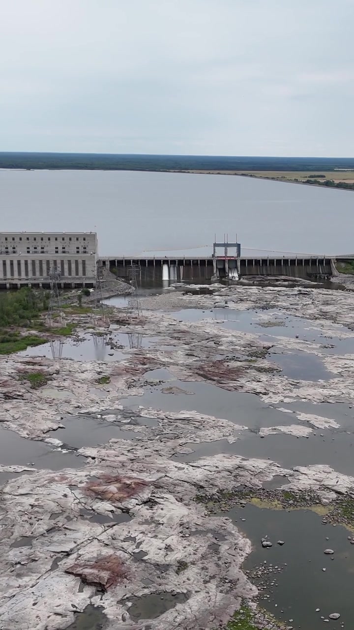 Ripresa aerea verticale grandangolare con drone che vola in avanti da lontano verso la centrale elettrica Seven Sisters, mostrando rocce esposte dello Scudo Canadese, un canale di sfioro e un lago artificiale dietro la diga