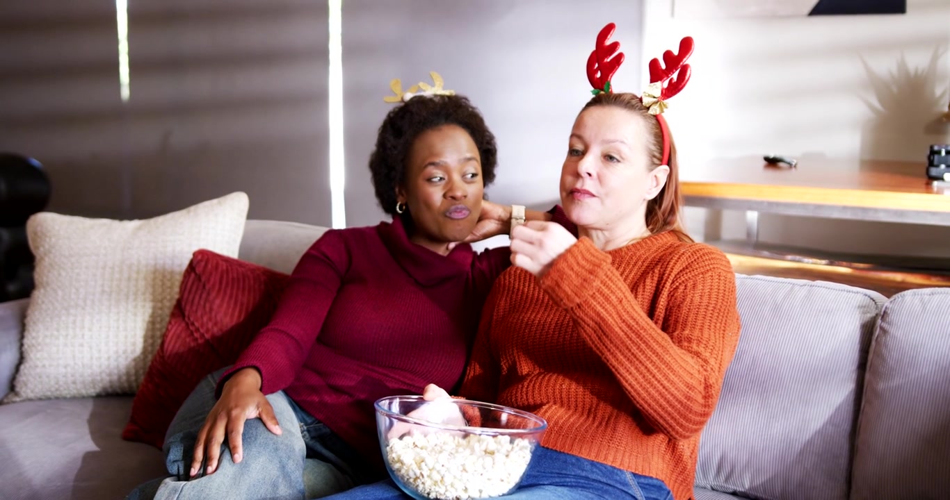 Venninner med ulik bakgrunn i oransje og burgunder gensere på sofaen deler popcorn mens julefilm spilles