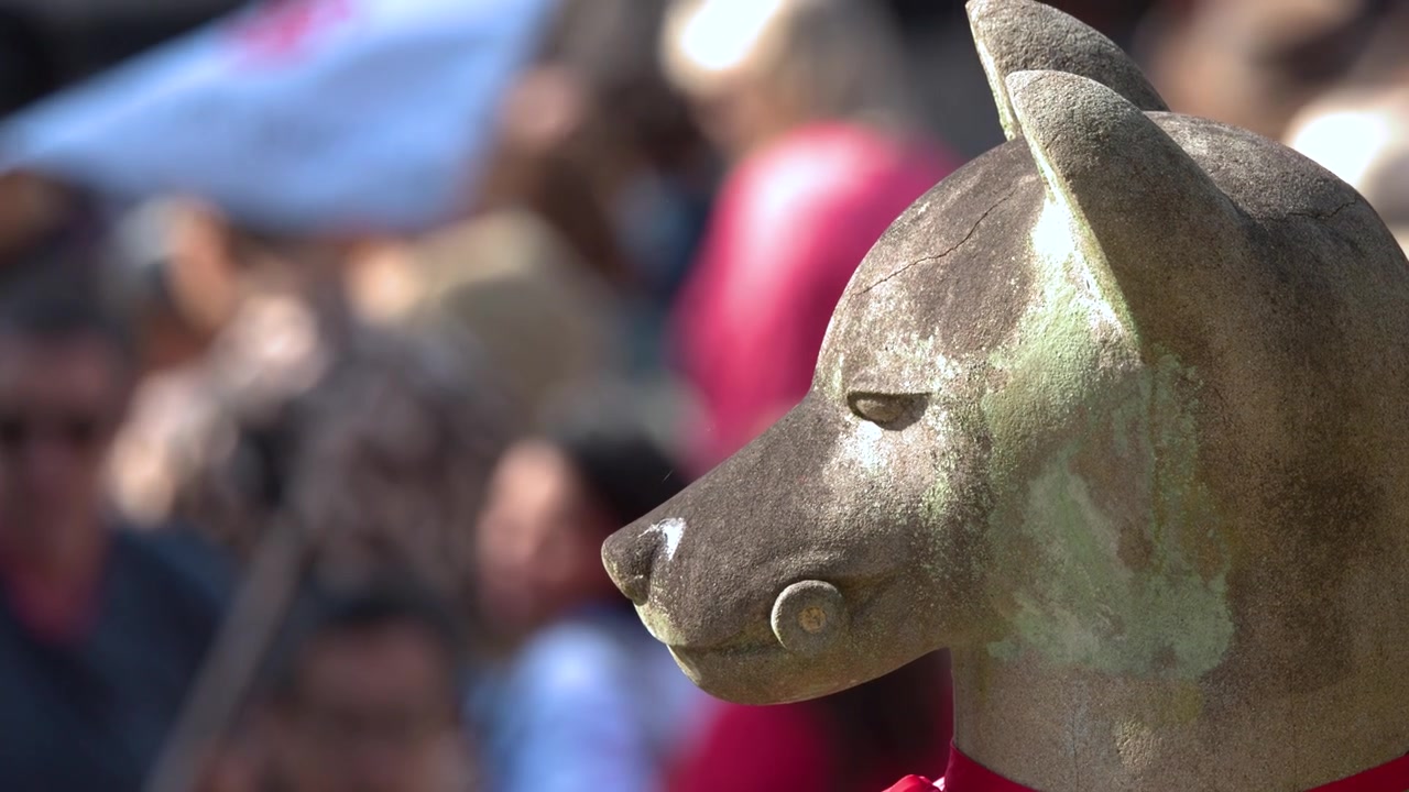 Primo piano di profilo di una statua di volpe in pietra con turisti sfocati che si muovono sullo sfondo.