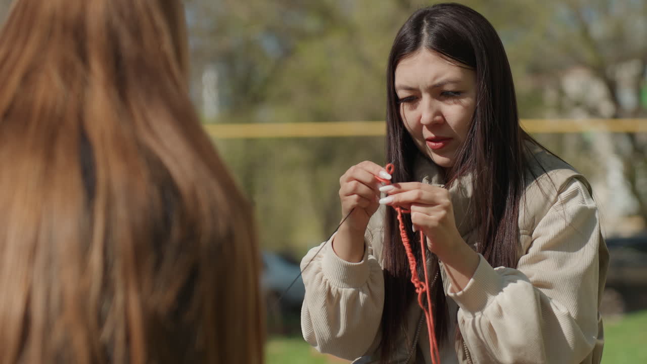 Instructor arreglando un tejido de punto, mentor guiando una puntada en un parque, profesor ajustando cuidadosamente un proyecto de hilo al aire libre, educador adulto dando instrucciones de tejido con paciencia en un entorno soleado