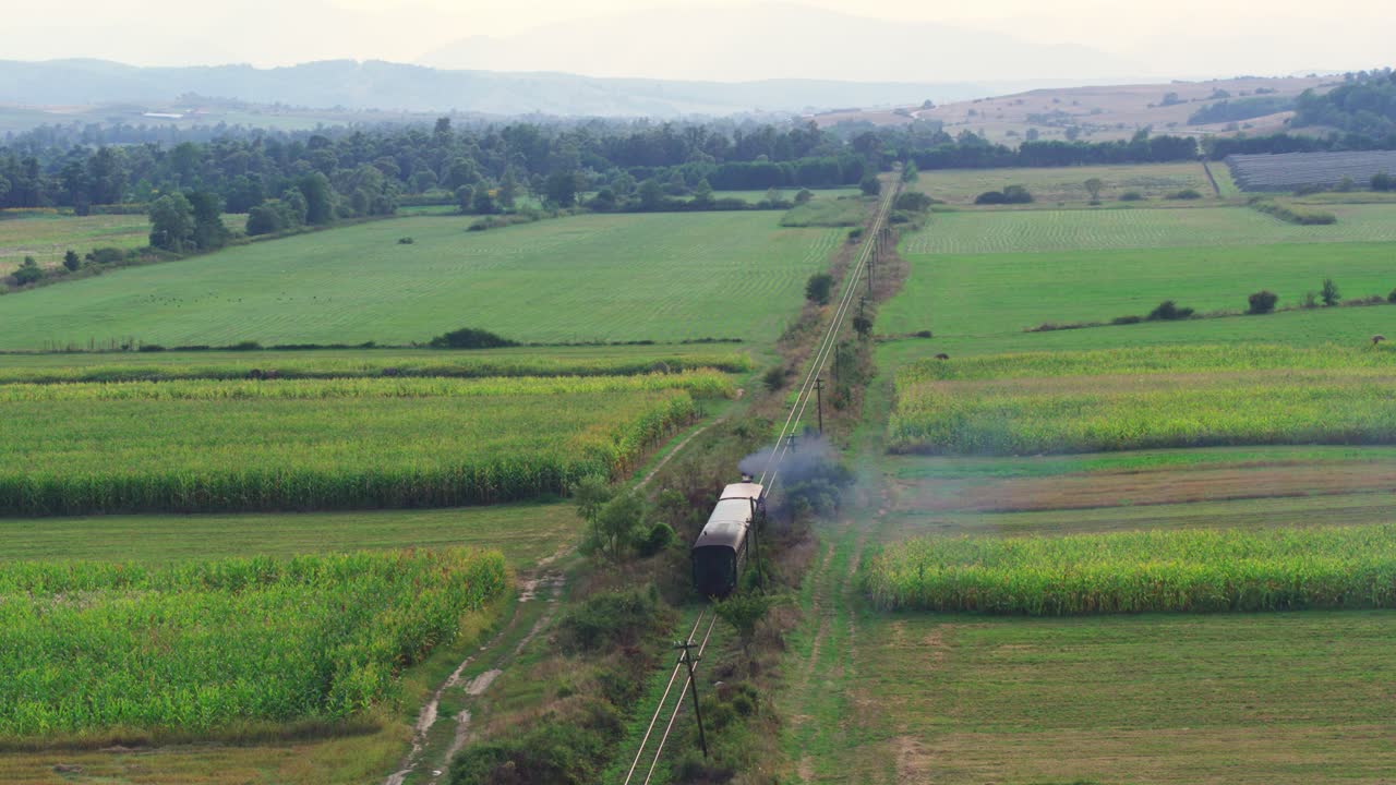 A beautiful aerial tracking shot follows a historic steam train on its journey. The locomotive travels through a vast, rural landscape, a patchwork of green and golden farm fields