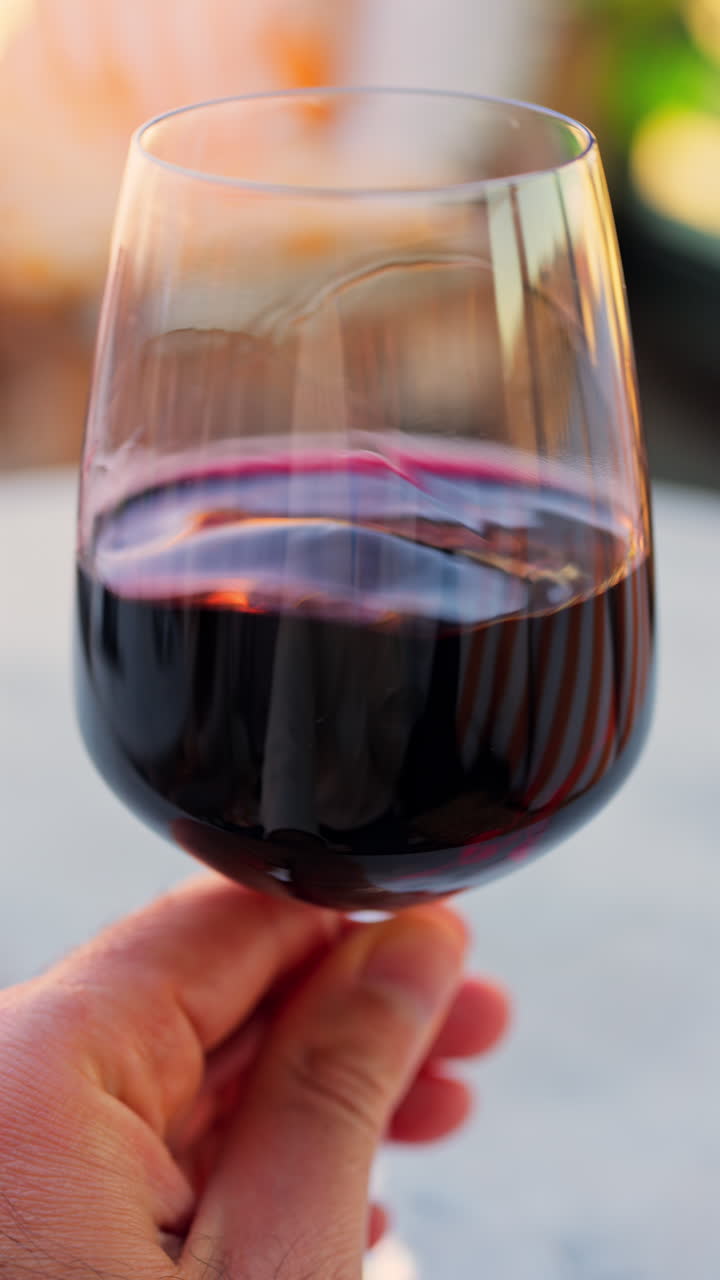 Close up of a woman's hand swirling a glass of red wine on a table at a terrace. Vertical