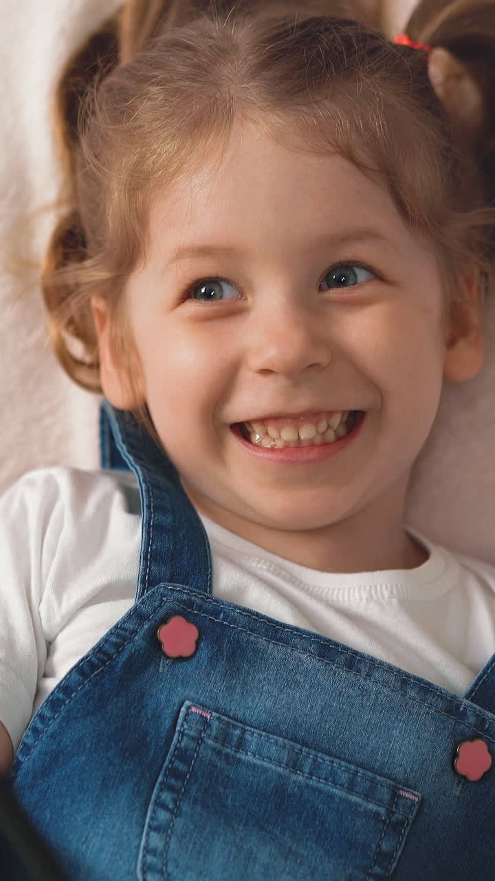 CU, High angle shot, Portrait: little girl, student child with long blond hair collected in pigtails, jeans sundress white T-shirt lies on floor laughs cutely, covers book hold pencil in hand closeup