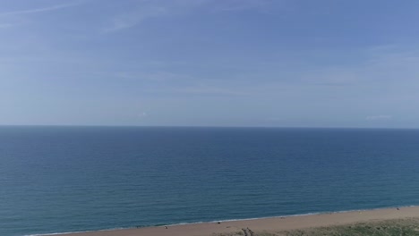 Aerial-tracking-out-to-gorgeous-blue-sky-and-sea-over-a-pebble-beach-with-a-lake-immediately-in-the-foreground