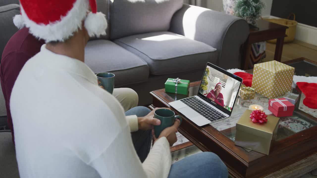 padre y hijo biraciales usando una computadora portátil para una videollamada de navidad con una mujer feliz en la pantalla