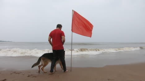 perro pastor alemán en la playa con su dueño cerca de una bandera roja | amistad entre humanos y animales | bandera de alerta alta en la playa