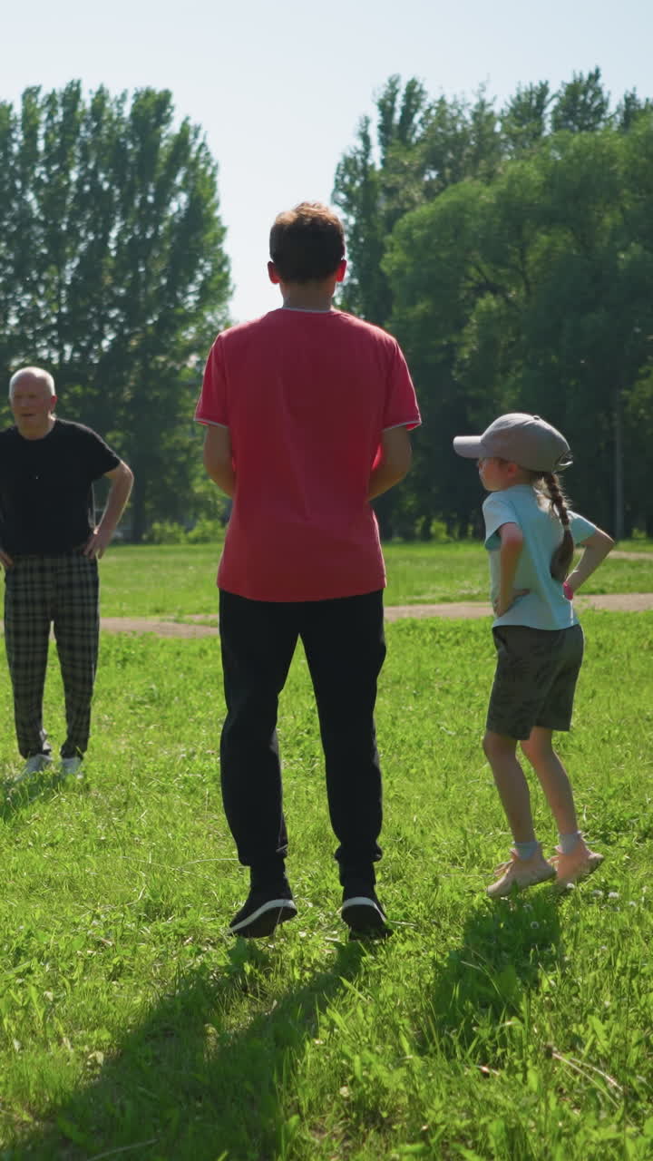 una familia está jugando al aire libre, el abuelo lleva a sus nietos, que están saltando con él, un niño sostiene una pelota, mientras que su padre lanza otra pelota hacia ellos
