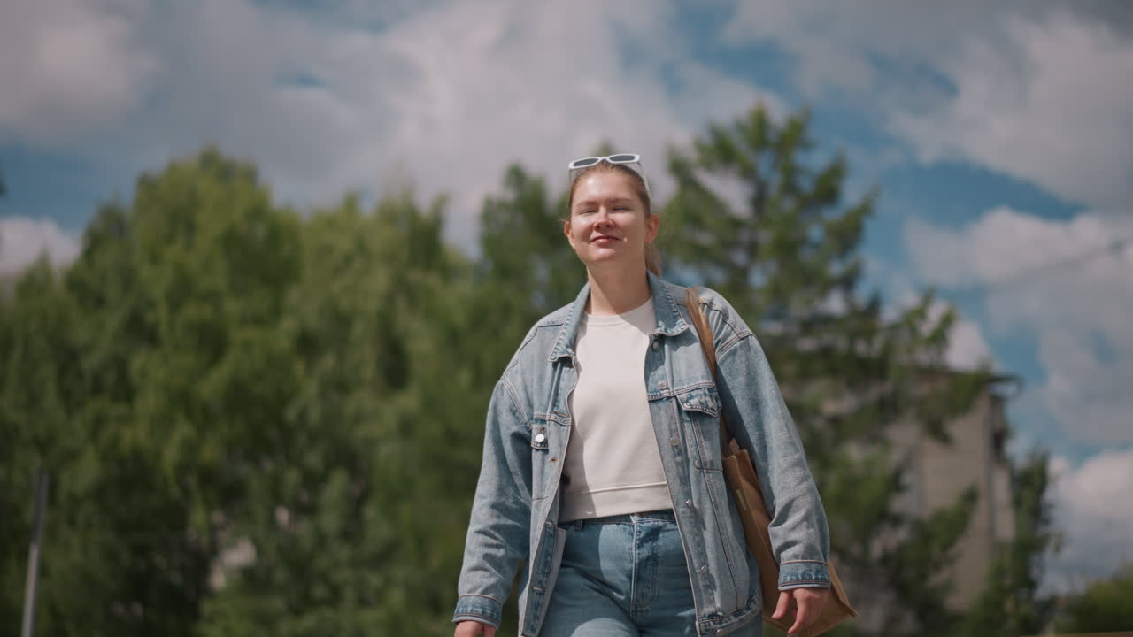 Joven mujer caucásica caminando bajo el cielo, chaqueta y jeans de mezclilla, bolso de hombro, nubes brillantes y árboles verdes de fondo, sonrisa confiada y paso ligero, ambiente de viaje de verano, estilo de vida espontáneo.