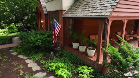 Aerial-shot-of-quaint-American-home-displaying-American-flag-above-beautiful-garden-and-landscaping-foliage