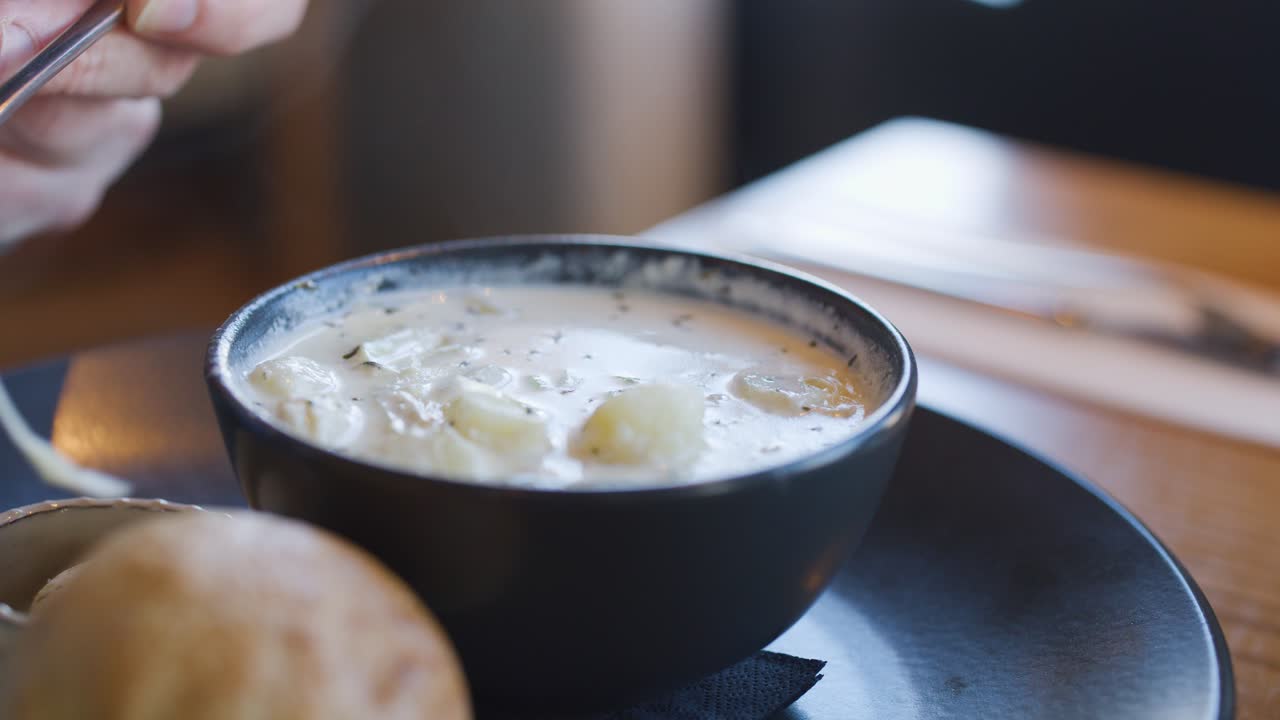 A hand lifts a steaming spoonful of creamy Cullen skink soup from a black bowl, with natural daylight and shallow depth of field enhancing the inviting winter atmosphere