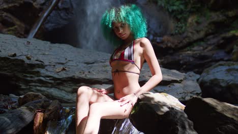 young bikini girl with green hair sits at the base of a waterfall in the caribbean