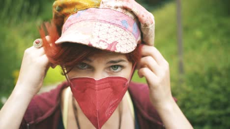 close-up of a young red haired woman puts on a medical red ffp2 mask with green nature background in slow motion