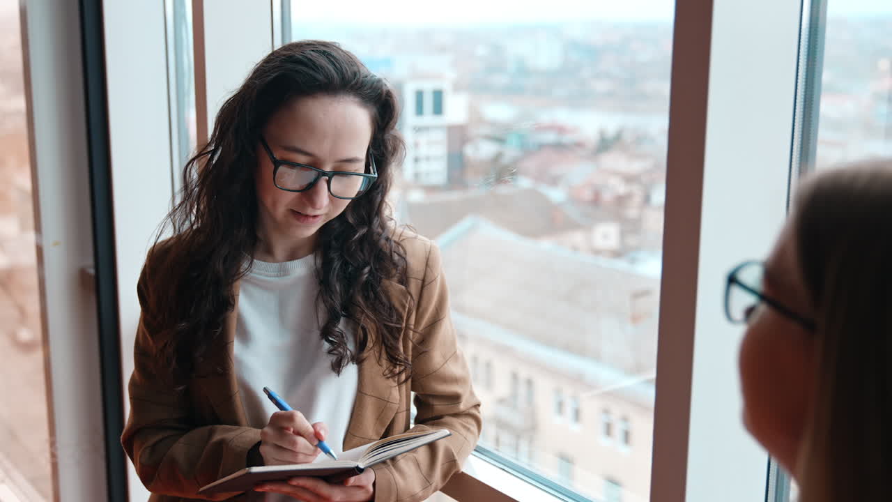 Secretary taking notes into paper notebook. Pretty brunette stands at the window, listens to her manager and writes down. View from top.