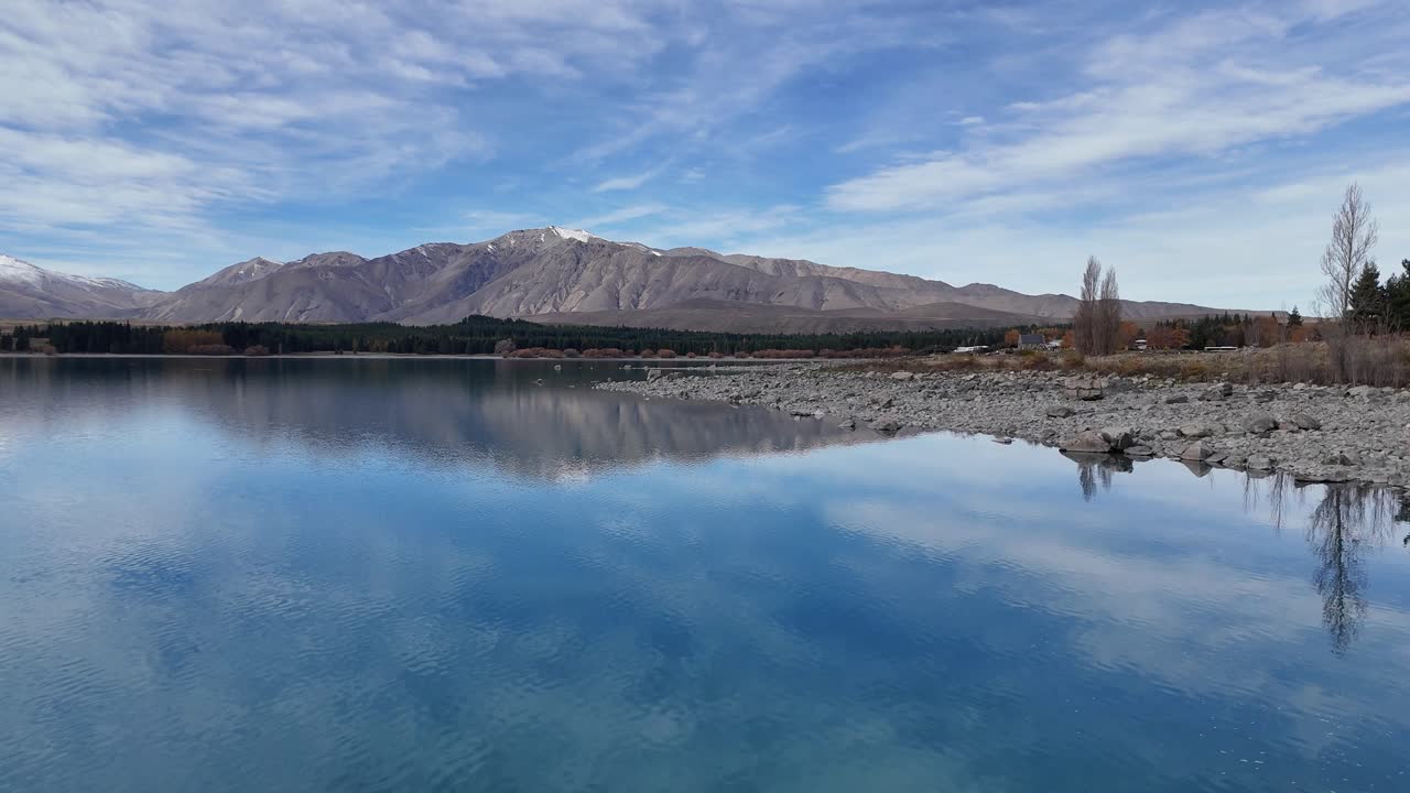 Aerial footage of Lake Tekapo captures tranquil waters, rocky shores, and distant mountains under a clear autumn sky