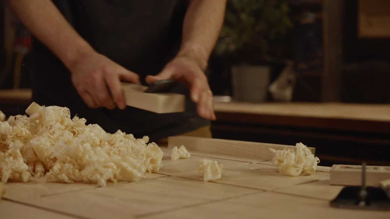 Primo piano delle mani di un artigiano che pialla un'asse di legno con trucioli sparsi sul banco da lavoro, a simboleggiare artigianato, precisione e lavorazione tradizionale del legno