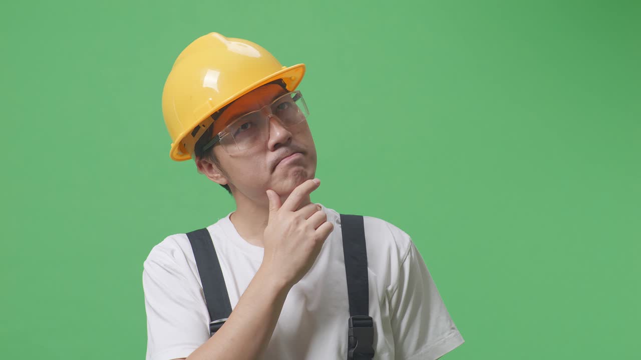 안경과 안전 헬을 착용한 아시아 남성 노동자가 생각하고 주위를 둘러보고 녹색 화면 배경 스튜디오에 서있는 동안 지표손가락을 들어 올립니다.