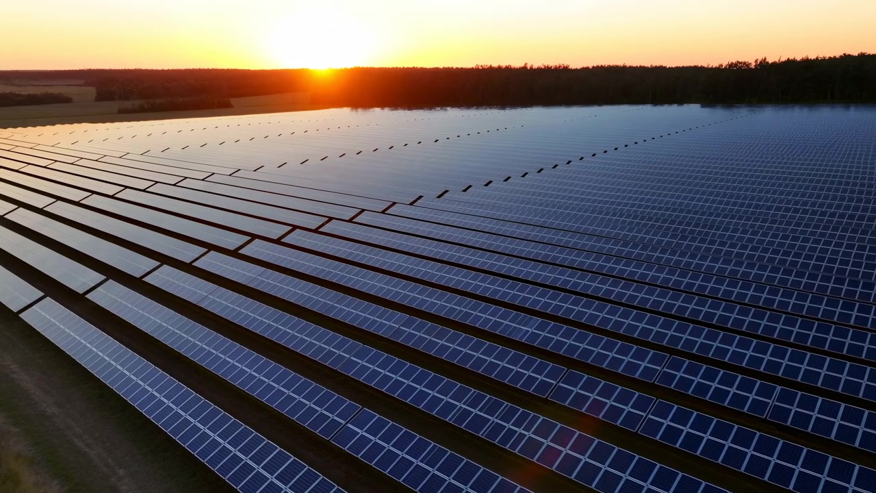 Vista angolata dall'alto su una vasta fattoria solare al tramonto, che mostra file diagonali di pannelli fotovoltaici blu scuro su strutture metalliche con stretti sentieri a terra.