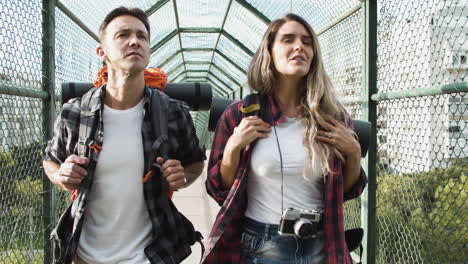Couple-of-travelers-with-backpacks-walking-on-bridge-from-city