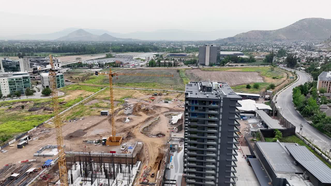 Forward drone aerial of construction site with yellow crane, foundation excavation, modern high-rise tower, and mountains valley backdrop