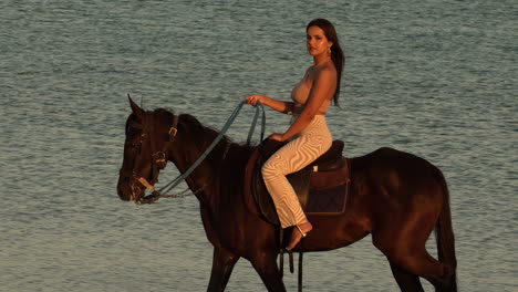 Hermosa-Joven-Elegante-Montando-Un-Caballo-Negro,-Con-El-Mar-Al-Fondo