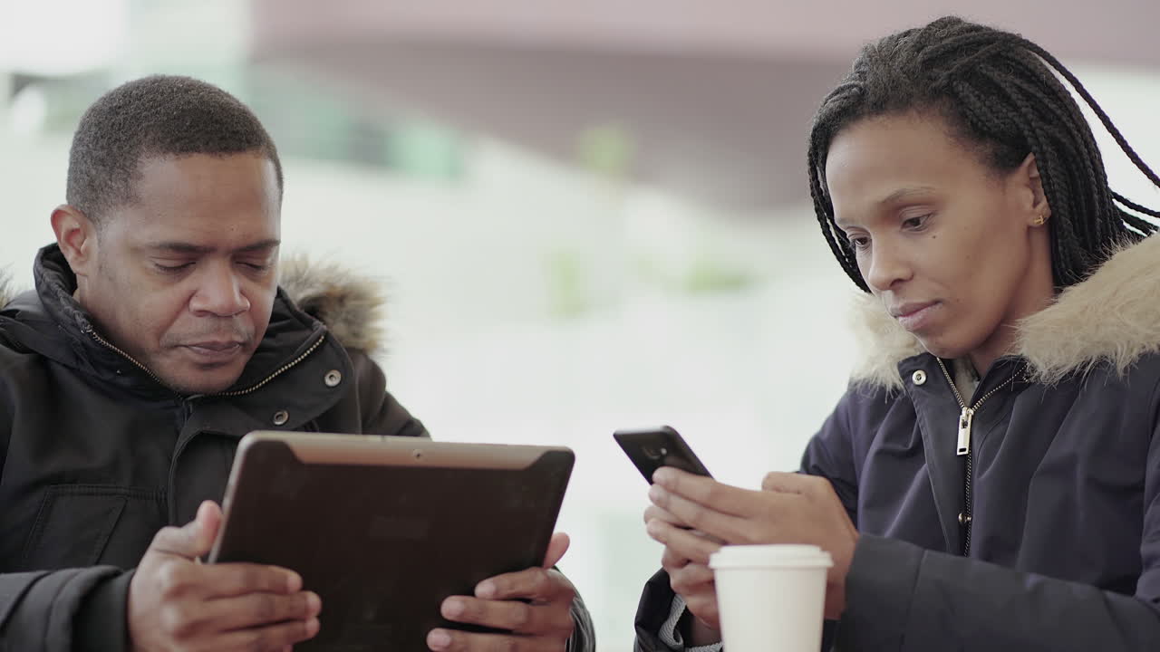 Afro-American young girl with braids googling on phone and Afro-American middle-aged man in jacket with fur hood using tablet