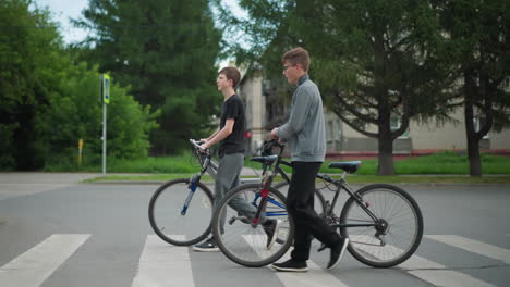 two friends walking side by side with their bicycles across a pedestrian crossing, a blurred view of three people walking in the background, with trees and residential buildings visible