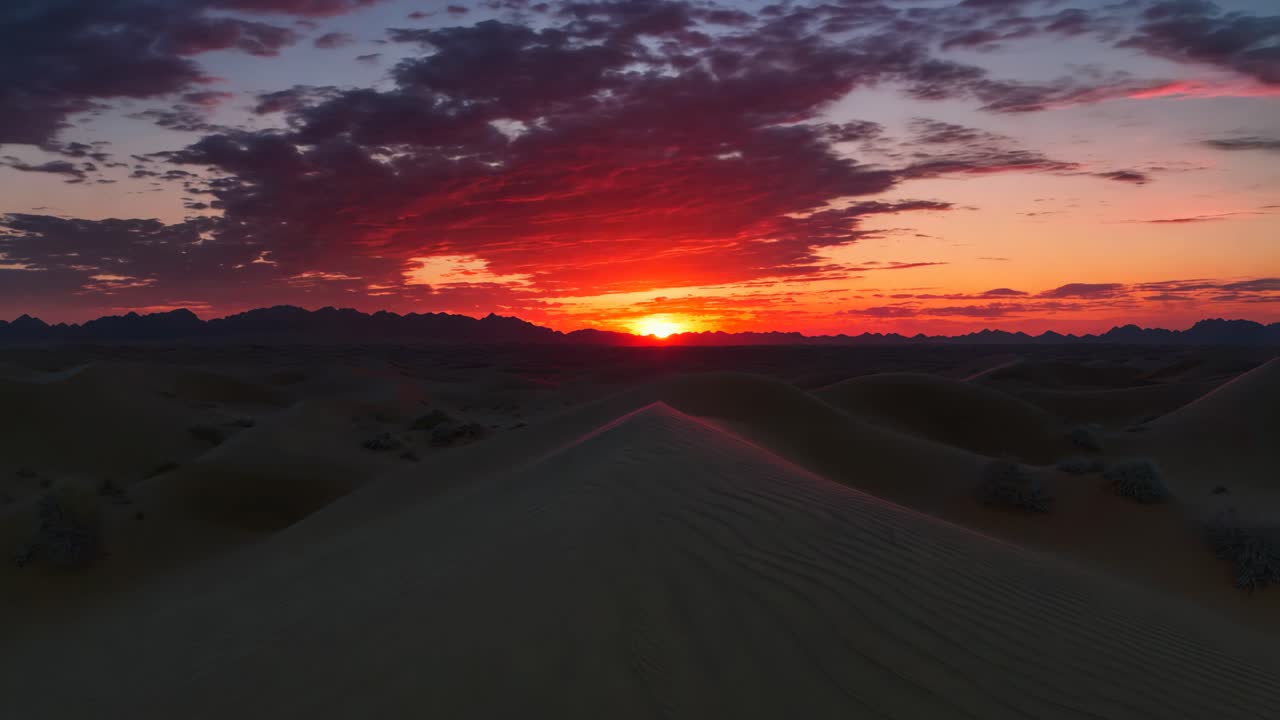 A desert landscape with a red sun in the sky. The sun is setting and the sky is filled with clouds