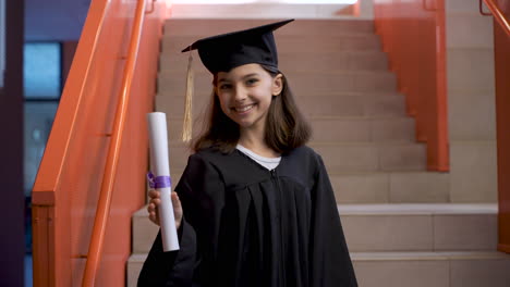 portrait of a happy preschool female student in cap and gown running down the stairs, holding graduation diploma and looking at the camera