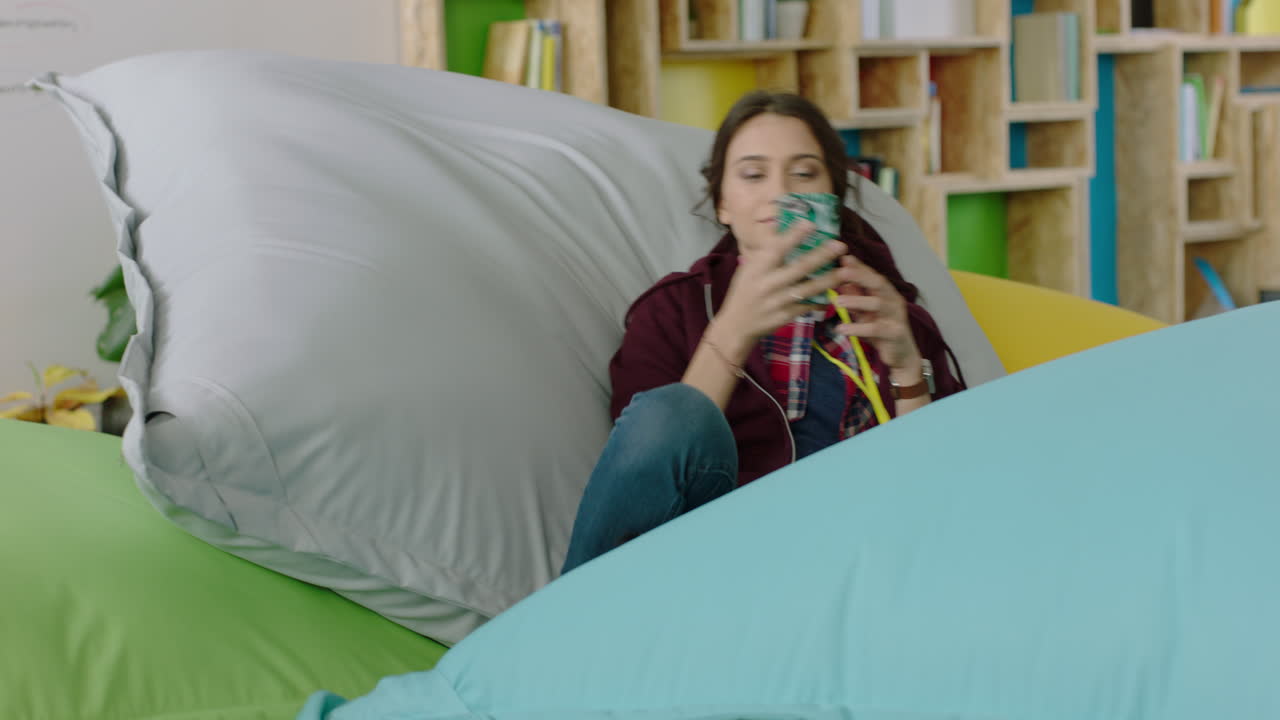 estudiante feliz usando un teléfono inteligente enviando mensajes de texto navegando en línea mensajes de redes sociales saltando sobre almohadas coloridas relajándose en una oficina moderna de moda