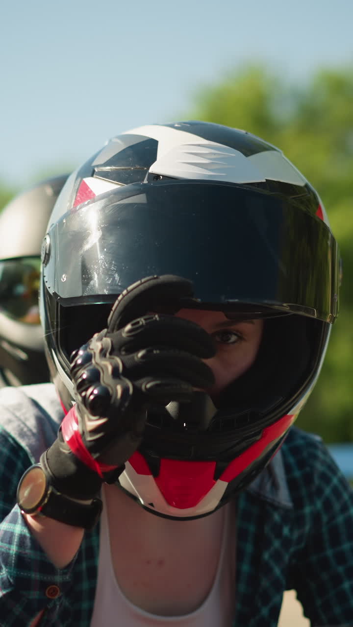 Two friends ride a red power bike, both wearing helmets, with the rider closing her visor, the rider wears black gloves and a wristwatch, a car is visible behind them
