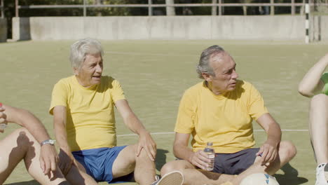 tired senior football players sitting in circle on pitch, talking together and drinking water